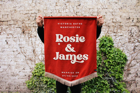 Terracotta velvet wedding banner with "rosie & james" names, venue, and date in small text. Person holds it up against an ivy brick background.